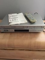 Front view of silver JVC DVD player and VHS VCR combo unit on wooden surface with remote control and instruction manual on top.