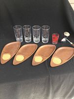 Four teak plates with cork inserts placed in a row in front, four clear hiball glasses with bowling ball embossing in the back, one small red shot glass, and metal measuring spoon with faux bone handle at side.