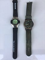 Two men's wristwatches side by side on a white background, one black Reebok digital watch and one analog watch with green fabric strap.