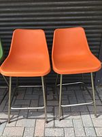 Front view of two orange counter height stools with molded plastic seats and metal legs showing overall condition and color.