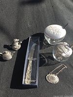 Two glass jars with ornate pewter lids and matching spoons, pewter cheese cutter in original box, pewter red wine label, and two small pewter teapot decorations placed on fabric background.