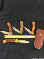Four folding lock blade knives laid out on black fabric, each with wood handles and brass-colored bolsters; an adjacent leather knife pouch is visible. A tape measure shows blades approximately 3 to 3.5 inches long.