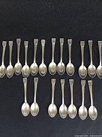 19 pewter spoons laid out showing handles with Canadian provincial coats of arms and other crests, photographed on black fabric background.