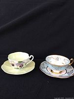 Two bone china teacups with matching saucers displayed side by side, showing the Shelley Pansy pattern on pale yellow and the Woodlands blue and gold pattern.