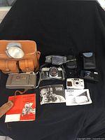 Overview of all cameras including old Polaroid Land Camera with case and manual, Canon EX Auto camera with leather case and instruction booklet, Pentax Espio 145M Super camera with case and manuals, plus additional accessories like flash units.