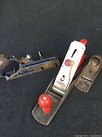 Three vintage hand planers laid on a black surface. Featured are the Este No 5 with red knobs, blue Record No 078 worn planer, and small Stanley No 220.