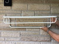 Side view of white wall-mounted metal wire shelf held horizontally by a person, showing tubular frame structure and mounting points.
