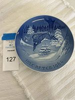 Front view of the 7 inch B&G porcelain plate showing a winter scene with a hare jumping among snow-covered fir trees, with the text 'JULE AFTEN 1964' at the bottom, photographed on a white cloth.