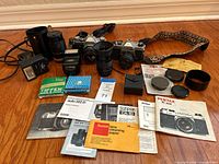Full overview of vintage camera equipment including Canon AT-1, Pentax K1000, lenses, filters, flashes, and accessories arranged on wooden floor.