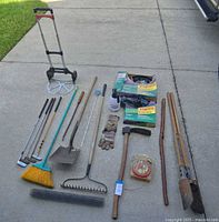 Full lot overview with all items laid out on concrete driveway showing hand truck, rake, shovel, broom, golf clubs, gloves, sprayer, fabric packages, axe, walking stick, stakes, wire roll.
