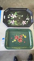 View of both metal trays on table, black tray with pink flowers and green tray with red and yellow flowers