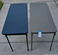 Two rectangular folding tables positioned side by side on pavement, showing black and gray colors.