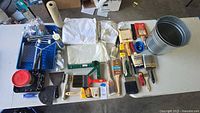 Full overview of all items laid out on table showing paint brushes, tray liner, metal bucket, roller frame and covers, caulking gun and tube, spreader, drop cloths, scraper, mixing sticks, paint pan and caulking tools.