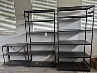 Three black metal storage shelves against wall; two tall units and one smaller unit to left. Shelves have five levels and matte black finish.