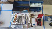 Image showing blue metal toolbox open and various assorted hand tools laid on white towel. Includes auger drill bits, hand saw, hacksaws, and wall hooks case.