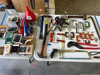 Photo showing hand tools laid out on table including hammers, pliers, clamps, utility knife, hacksaw, crowbar, and screws containers.