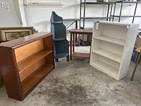 Four storage shelves including a wood shelf, a distressed blue corner shelf, a wood side table, and a white shelf on a concrete floor in warehouse setting.