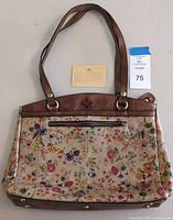 Front view of Patricia Nash ladies handbag showing floral canvas with brown leather trim, handles, and embossed leather patch with riveted hardware. Includes product information insert behind the handbag.
