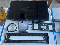 Samsung 28 inch LED TV model UN32J5205AF lying face up on a blue and white surface with included accessories around it: one HDMI cable, power cord, remote control, two metal wall mount brackets, and user manual papers.