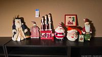 Full view of all 12 Santa Claus themed holiday decor items arranged on a black shelf, including tabletop figurines, mugs, and framed art piece.
