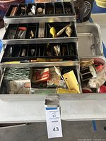 Metal tackle box with open compartments showing assorted fishing lures, hooks, lines, and accessories inside.