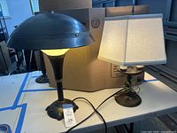 Photo showing two vintage lamps, one with black metal mushroom shade and one with hexagonal fabric shade, on a table