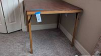 Front and angled view of vintage brown folding wooden card table set against a corner with carpet floor and beige walls.
