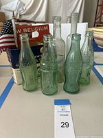 Four vintage Coca-Cola bottles and one vintage soda water bottle placed in front of a New Belgium Fat Tire package on a table with a small American flag