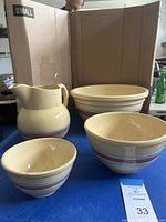 Three nesting ceramic bowls with decorative pastel horizontal stripes and a matching ceramic pitcher, all displayed on a blue surface in front of a cardboard box.