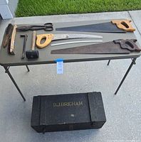 Image showing vintage hand saws, scissors, hammer, brush, hook tool on table with black wooden storage trunk beneath