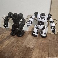 Two WowWee Robosapian robot toys standing on a wood floor, one black and one white with black details.