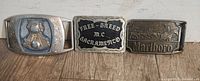 Three brass and metal western themed belt buckles side by side on wooden surface.