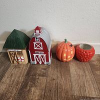 Four kitchen-themed decorative items: green roof hut, red barn cookie jar, ceramic pumpkin, and red textured bowl, all on wooden floor.