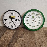 Photo showing both wall clocks side by side on a floor against a wall. John Deere clock on the right with green frame and tractors; Hertz clock on the left with black frame and equipment images.