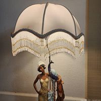 Porcelain table lamp lit, showcasing lady figurine holding a peacock and decorative beaded fringe shade.