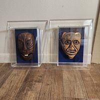 Two Moyang Mah Meri tribal masks side by side, each in its own clear acrylic display case, both resting upright on floor against wall.