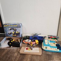 Lot of toys including a robot arm kit, various toy vehicles, wooden animal puzzle pieces, and a blue toy boat on a wooden floor