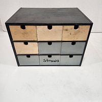 Front view of the wooden organizer box showing nine drawers of different colors and one labeled 'Straws'.