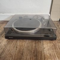 Front and side view of the Sony PS-T3 turntable with closed clear dust cover on worn wooden floor.