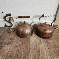 Photo showing two brass kettles and one brass dragon dinner bell placed on the floor against a wall.