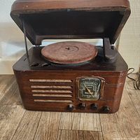 Front view of vintage wooden radio cabinet with open lid showing turntable felt pad, analog station dial, and four control knobs on front panel.