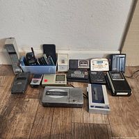 Photo showing the full collection of vintage phones, cellphones, and cassette recorders arranged in rows on a wooden floor against a wall.
