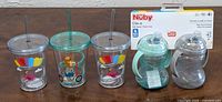 Five baby/kids' cups including 3 transparent Swirly Straw Tumblers with colorful animal designs (1 green with a teddy bear, 2 grey with flying pig-like characters) and 2 Nuby Clik-it Soft Spout Trainer Cups in green and grey with handles, all displayed on a white background.