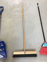 Wooden broom with dark bristles and blue snow shovel with black handle on gray floor.