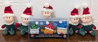 Five beanbag style Christmas elf plush toys with red hats, green outfits, white collar trim, standing in a row against a wall.