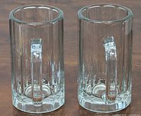 Two tall cylindrical glass beer mugs with thick panel cuts and wide handles placed side by side on a wooden surface, viewed from the front angle.