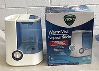 Front view of assembled Vicks Warm Mist Humidifier with blue 1 gallon water tank and original box next to it.