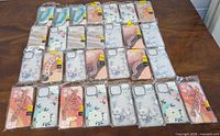 Photo showing 29 individually packaged iPhone phone cases laid out on a wooden surface. Cases feature floral and abstract design patterns mostly in pastel colors.
