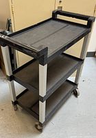 Side view of the black 3-tier Rubbermaid rolling cart showing three flat shelving layers with metal supports and caster wheels.