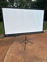 Movie projector screen set up outdoors showing the full 100-inch white screen with black border on tripod stand.